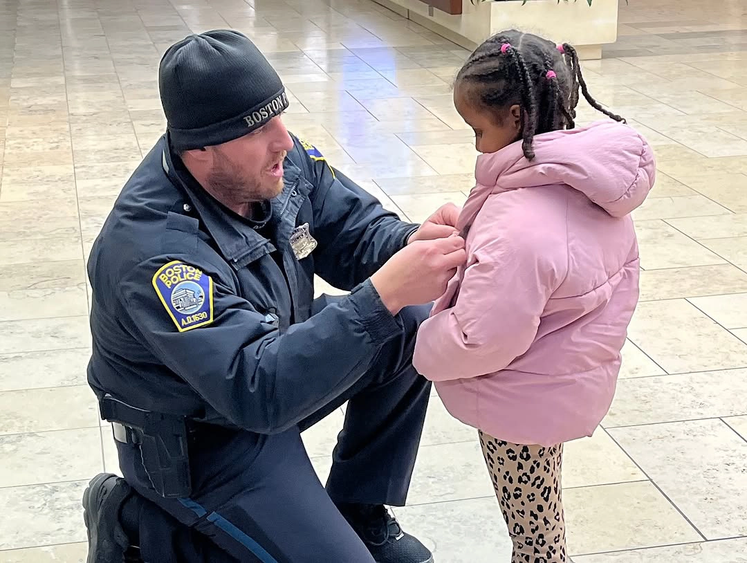 Boston Police officer kneeling to give a junior badge to a young child in a pink jacket at Prudential Mall