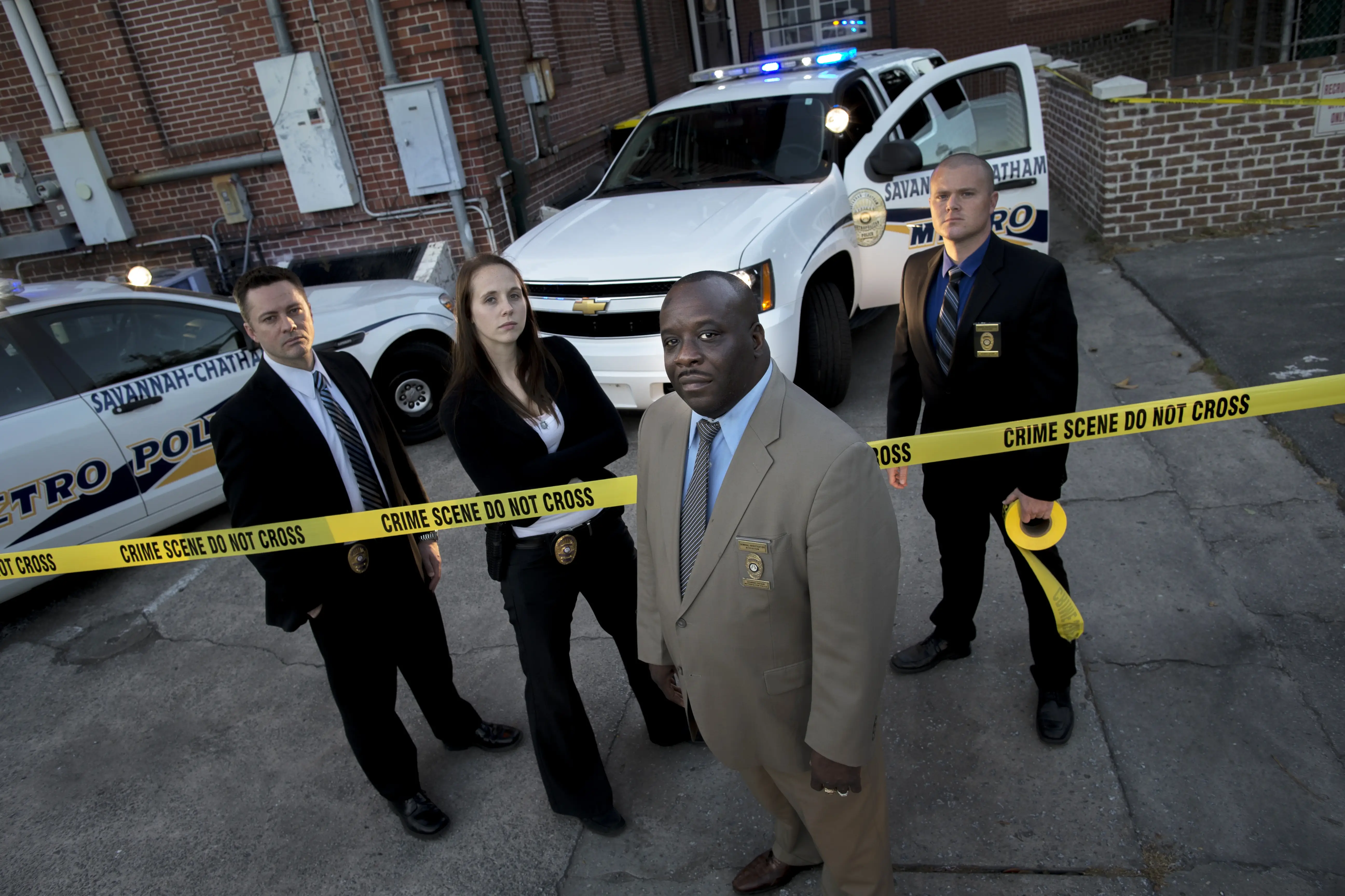 Homicide detectives standing at a crime scene with yellow tape and police vehicles in Savannah
