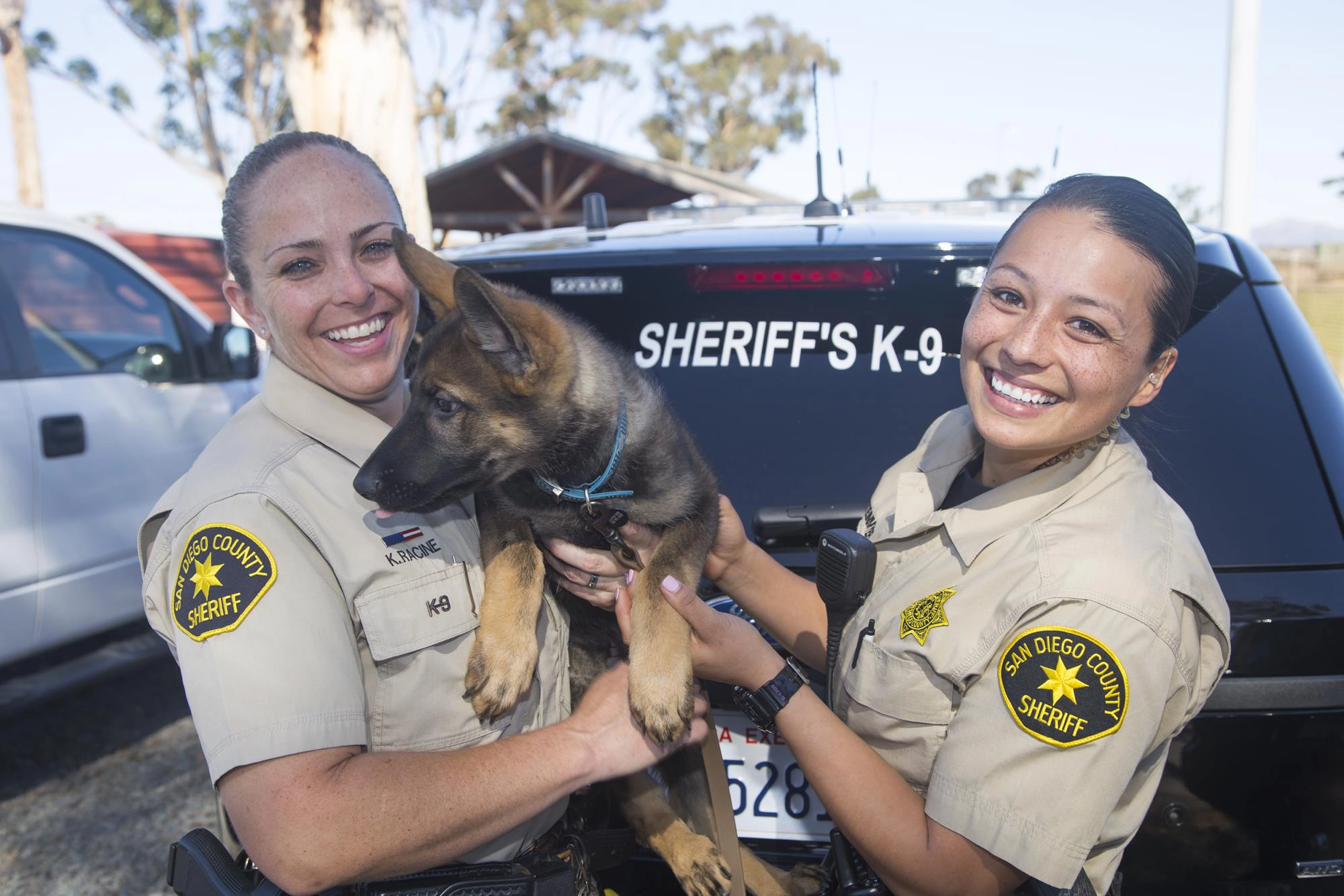 Two San Diego County Sheriff's deputies smiling with their K-9 partner in front of a Sheriff's K-9 vehicle